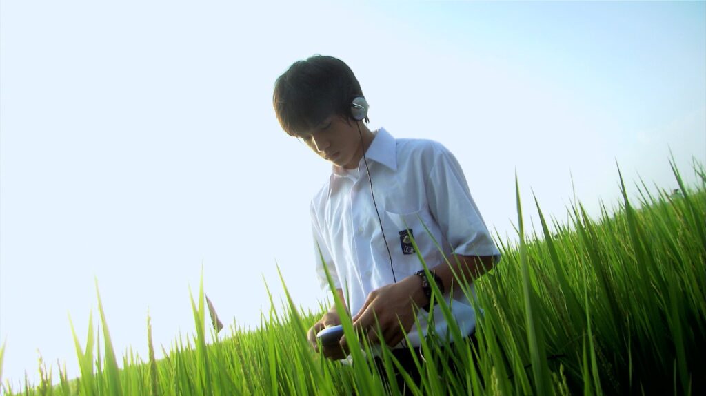 A student wearing headphones stands among rice fields in All About Lily Chou-Chou (2001), capturing youthful isolation.