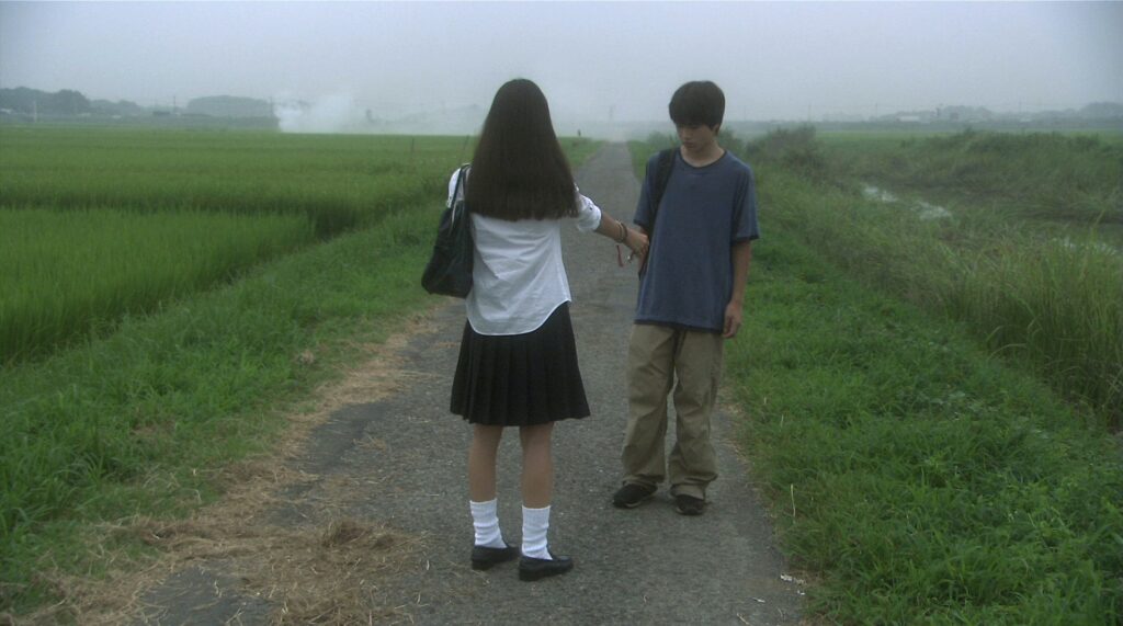 Two teenagers meet on a quiet country road in All About Lily Chou-Chou (2001), surrounded by the green fields of rural Japan.