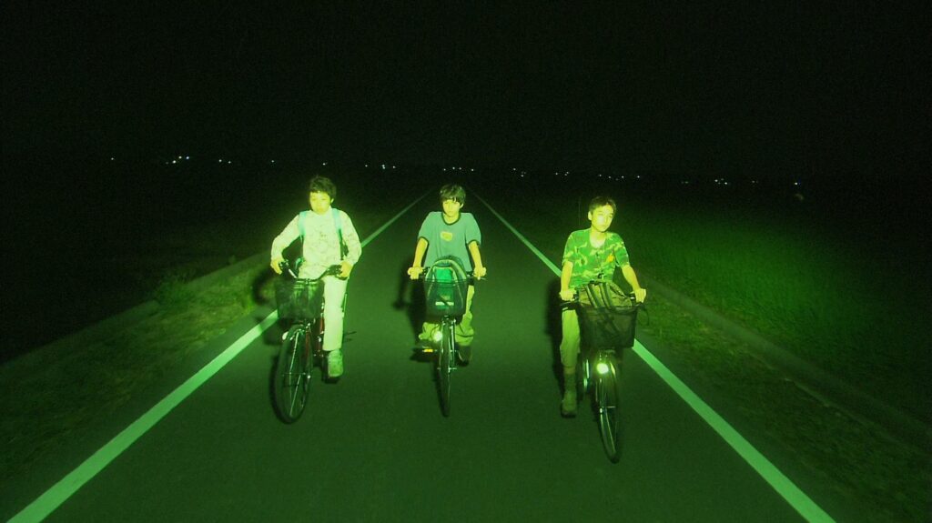 Three teenagers ride bicycles through the night in All About Lily Chou-Chou (2001), illuminated by eerie green light.
