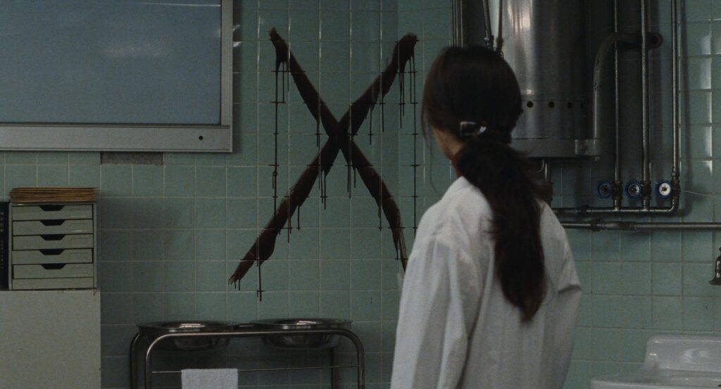 A woman faces a large X painted on a tiled wall in Cure (1997).