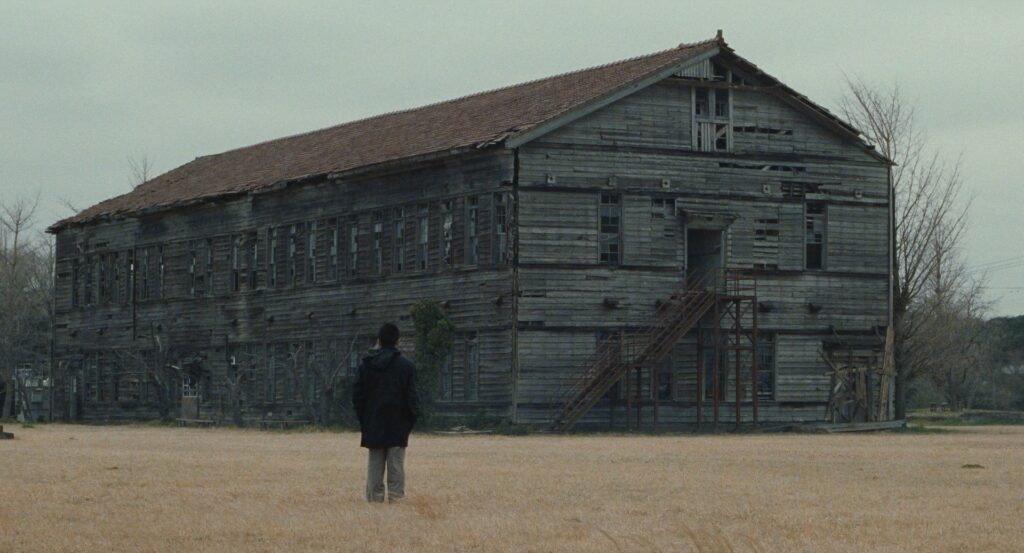 A man stands before an abandoned wooden building in Cure (1997).