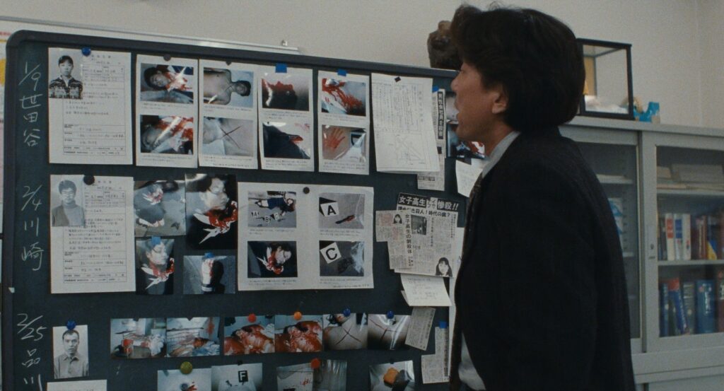 An investigator studies a case board filled with photographs in Cure (1997).