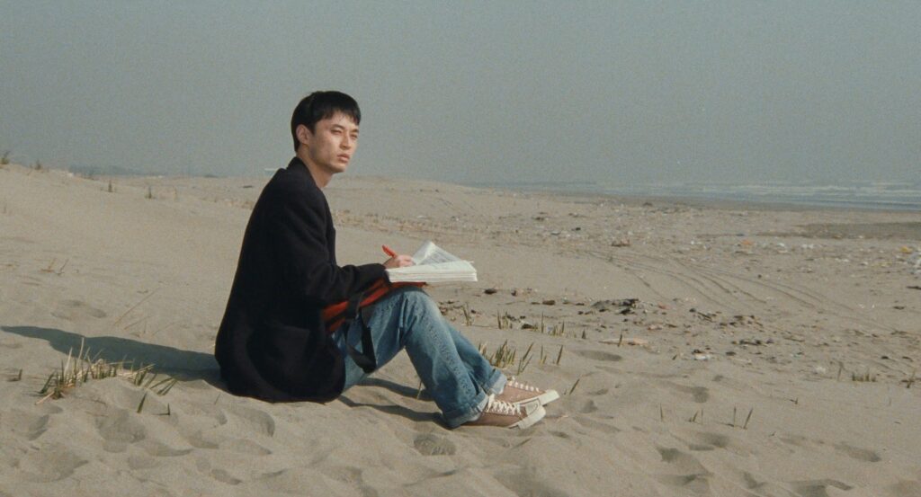 A young man sits alone on a beach in Cure (1997).