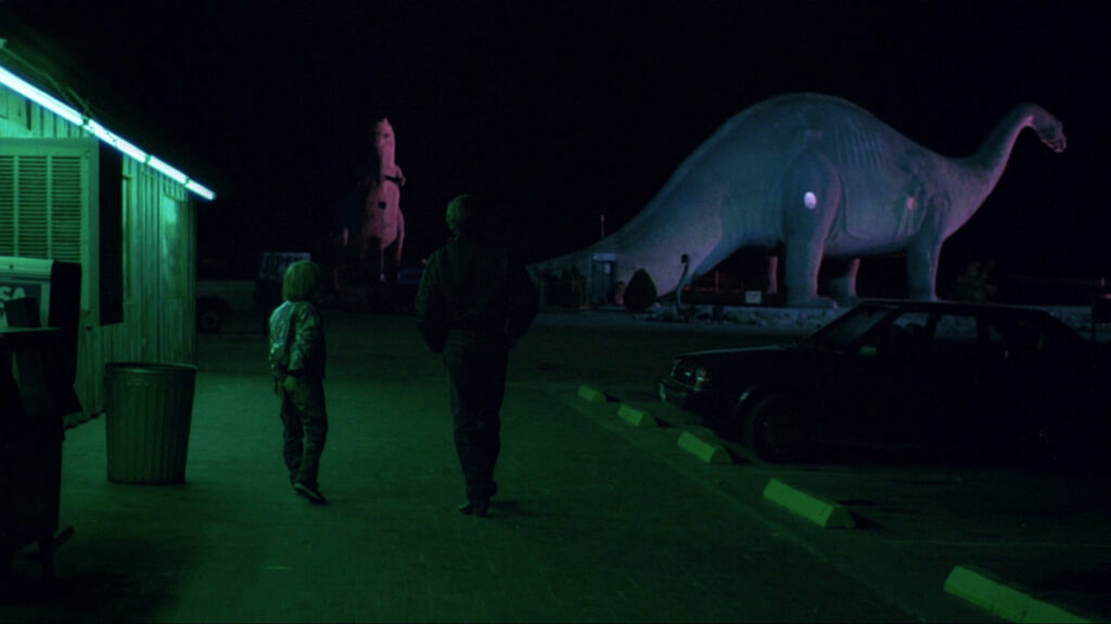 Travis and Hunter walk past a giant dinosaur attraction at night in Paris, Texas (1984).