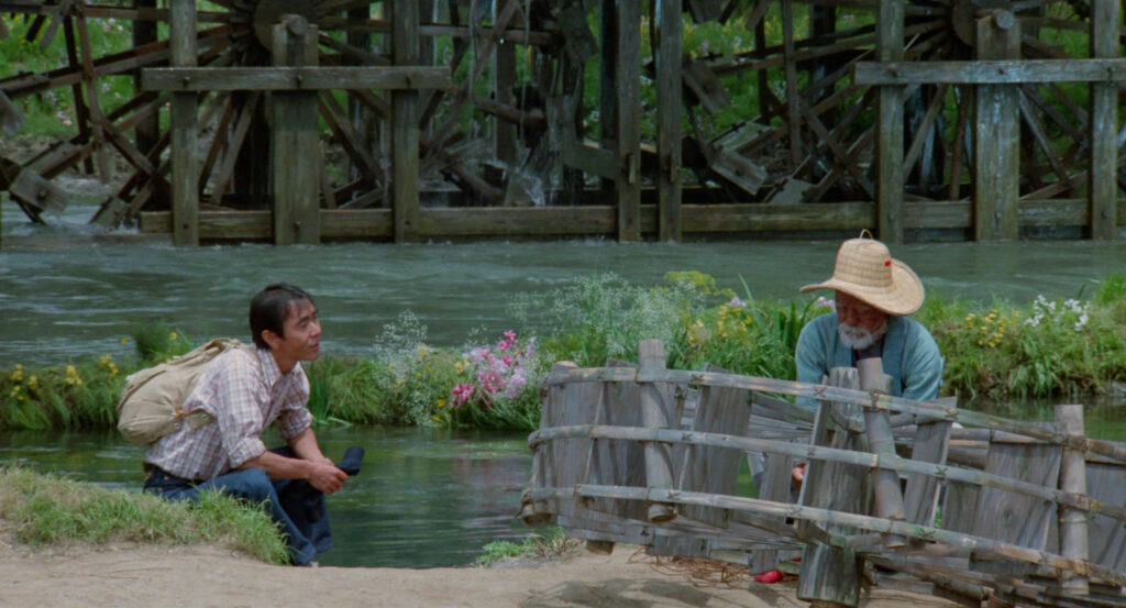 A traveler kneels beside an old man near a wooden waterwheel in Dreams (1990).