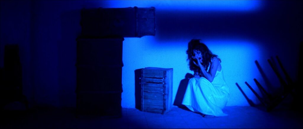 A frightened young woman crouches in a blue-lit room in Suspiria (1977).
