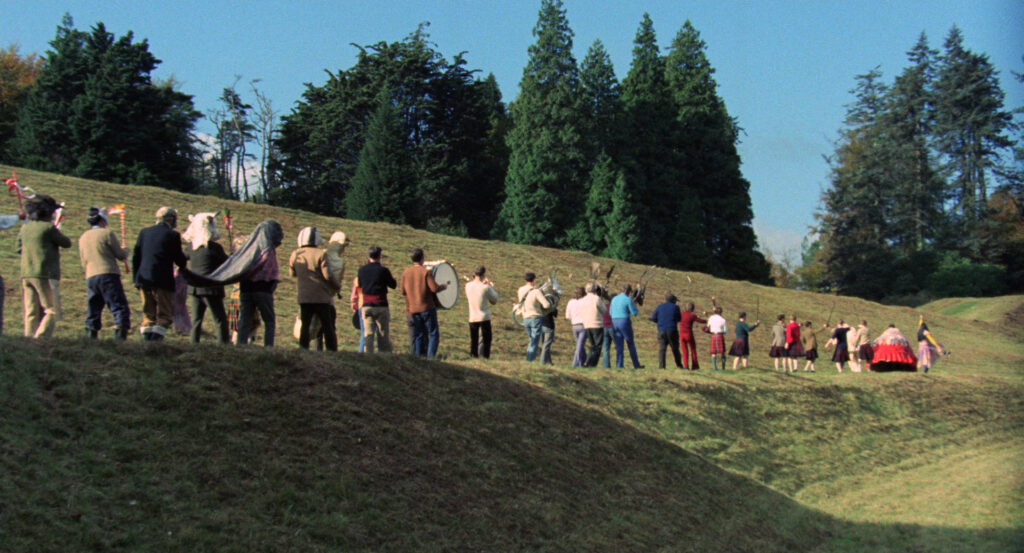 Villagers march across a grassy hillside during a festival procession in The Wicker Man (1973).