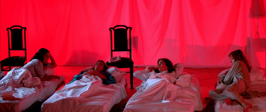Several students rest beneath glowing red curtains in a dormitory hall in Suspiria (1977).