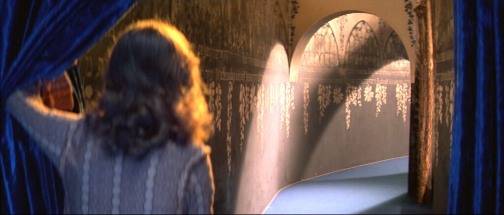 A young woman looks into a brightly lit corridor through blue curtains in Suspiria (1977).